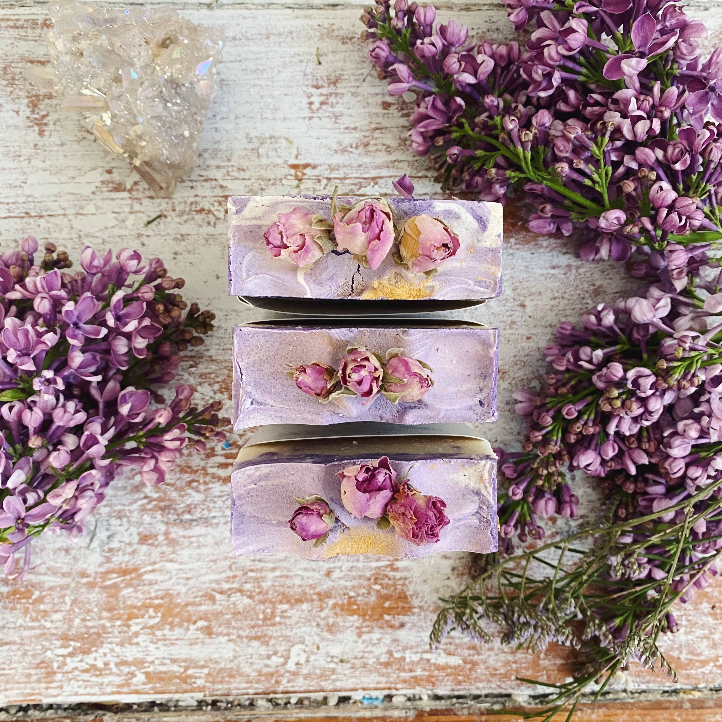 Three bars of soap with floral embedded ingredients on a wooden surface surrounded by lavender flowers.