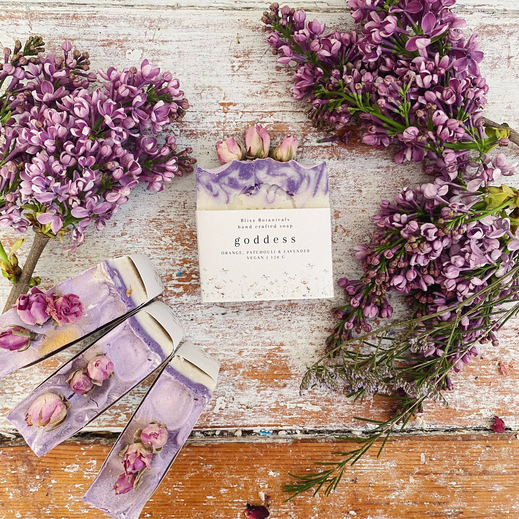 Bar of soap labeled 'goddess' with floral packaging on a rustic wooden surface with purple flowers.