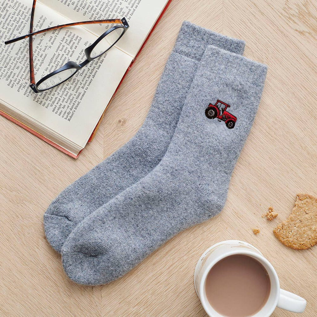 Gray socks with a red tractor design on a wooden surface with a book, glasses, and a mug.
