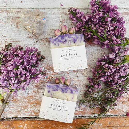 Two bars of soap labeled 'goddess' surrounded by purple flowers on a wooden surface.